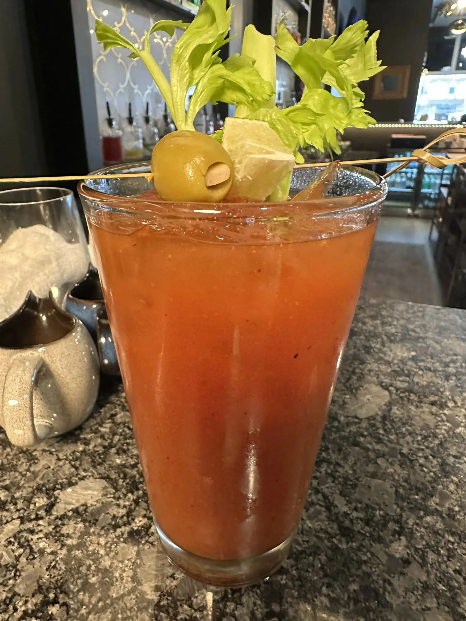 A tall Bloody Mary garnished with celery, olive and cheese on a skewer, resting on the bar at The Bar on Baldwin