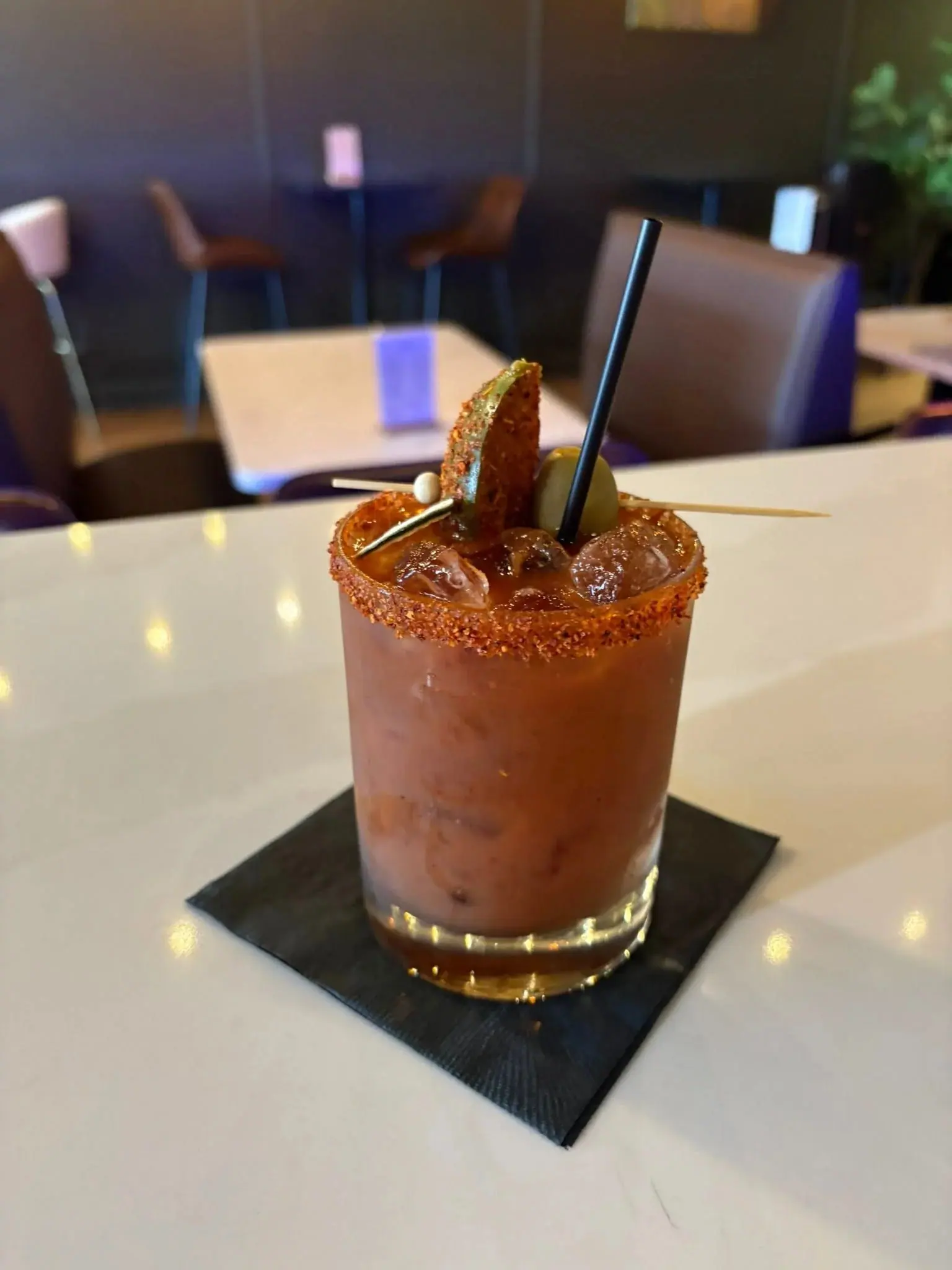 A Bloody Mary in a rocks glass with a chili-salt rim, olive and spiced lime, on a table in the dining room