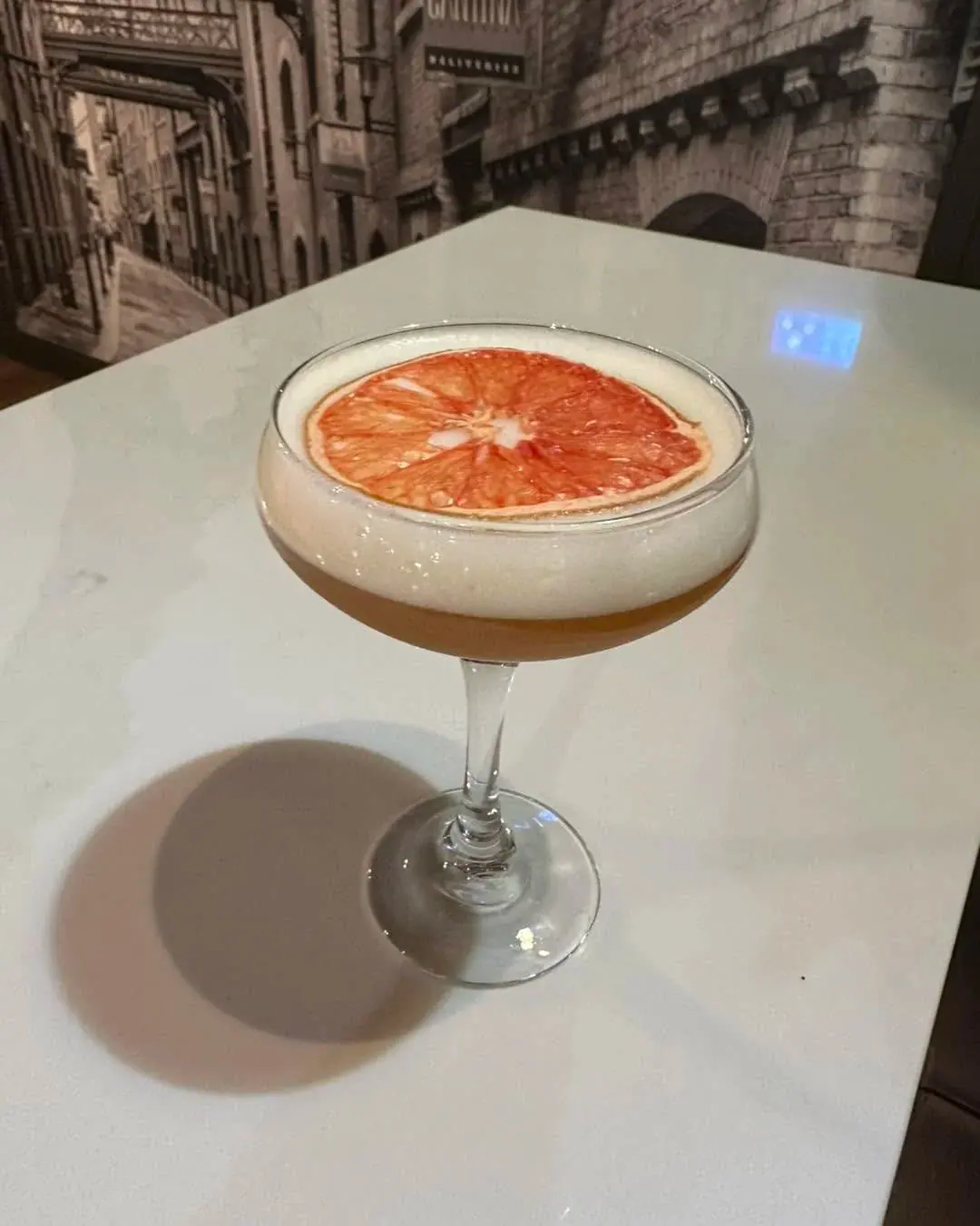 A coupe cocktail with a silky foam top and a dehydrated grapefruit wheel, on a pale marble table at The Bar on Baldwin